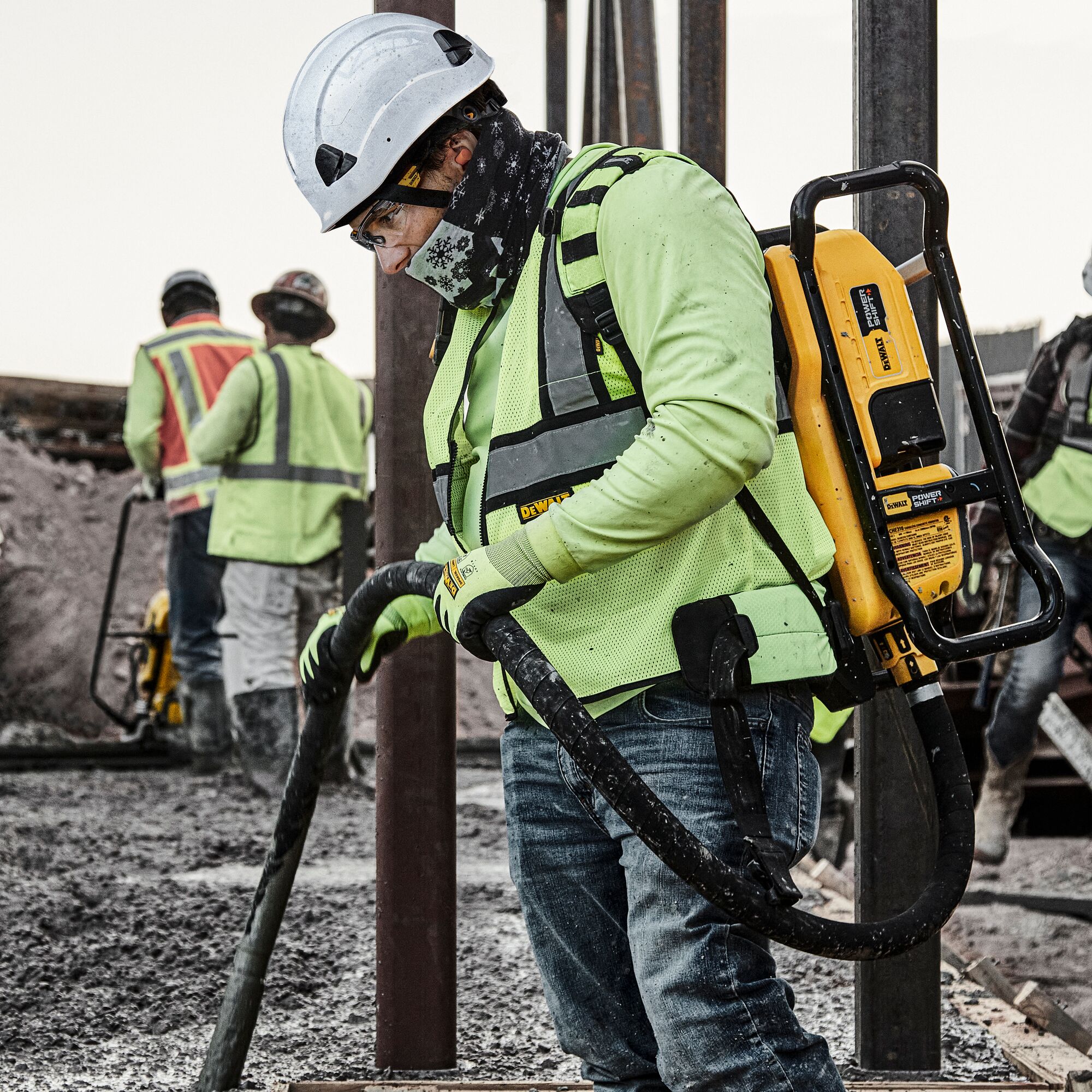 WORKERS ON A JOBSITE MAIN FOCUS IS MAN WITH DEWALT POWER TOOL WEARING WHITE TYPE II CHINSTRAPED HELMET, CLEAR SAFETY GLASSES, HI-VIS VEST AND GLOVES