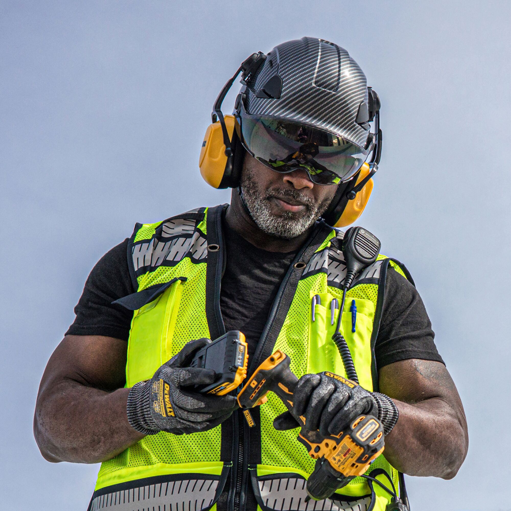 MAN WEARING A DEWALT HI-VIS SAFETY VEST, GLOVES, SLATE COLORED TYPE II VENTED HELMET, SMOKE VISOR AND CAPMOUNT EARMUFFS ATTACHING BATTERY TO A DRILL